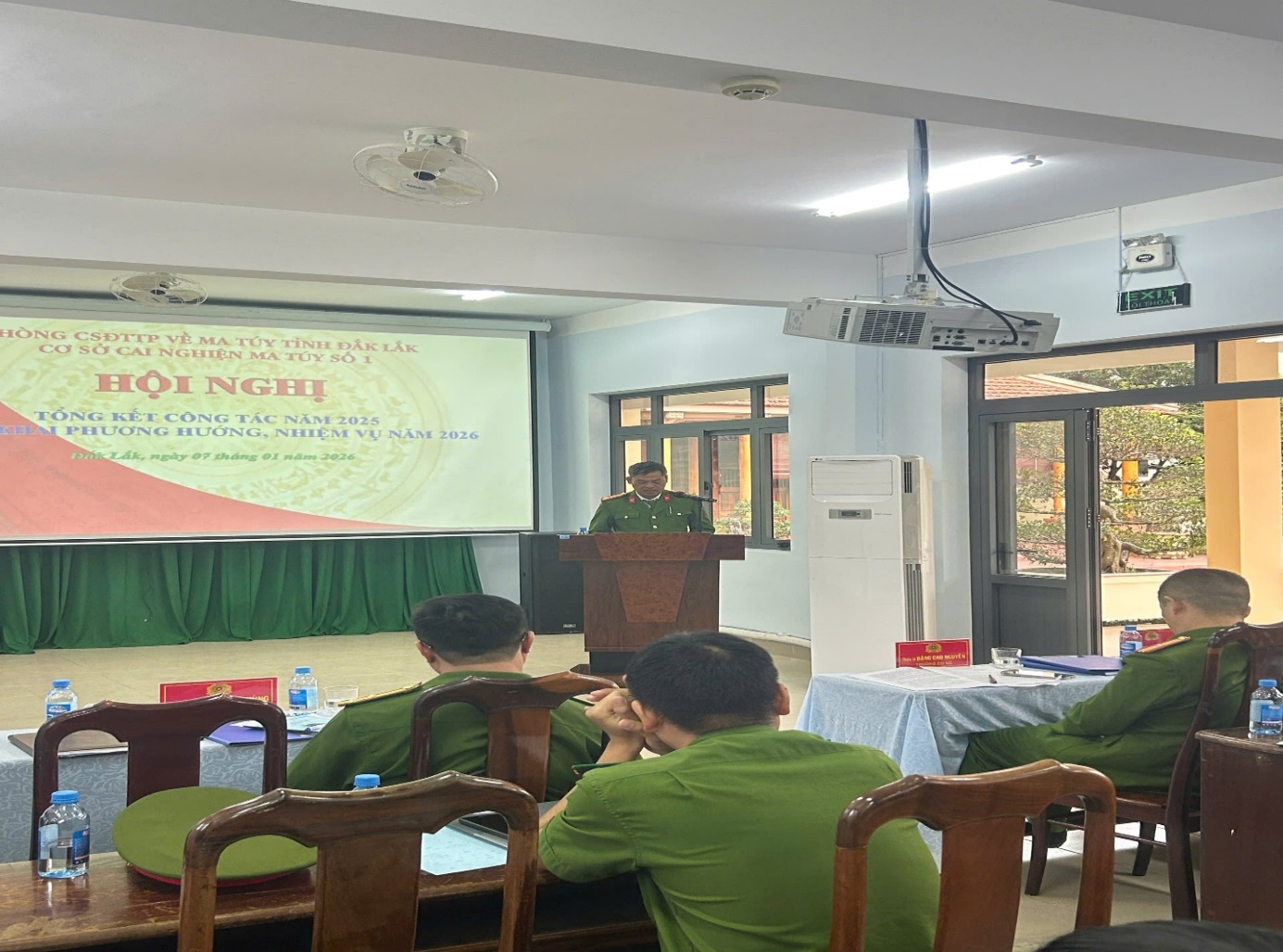 '''A person in green uniform standing in front of a podium with a projector screen
 Description automatically generated'''